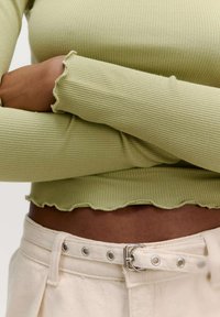 Geribbelde, lichtgroene longsleeve top met golvende zoom en manchetten, gecombineerd met crèmekleurige broek met een zilveren gesp en oogjes details.
