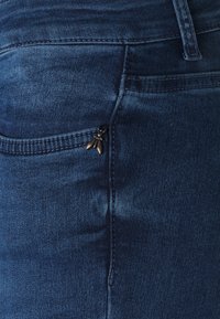 Jeans de mezclilla en azul oscuro, con un bolsillo con cremallera y un tirador de metal decorativo, costuras en contraste y una textura suave.