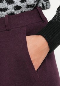Burgunderbukser har en glatt tekstur, sidelommer og detaljer i midjen. Hånden hviler i lommen, iført en svart stripete genser.