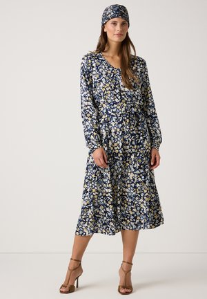 Blumenmuster-Kleid in Marineblau mit gelben und weißen Blumen, langen Ärmeln, taillierter Taille und ausgestelltem unteren Saum. Dazu wird ein passendes Kopftuch getragen.