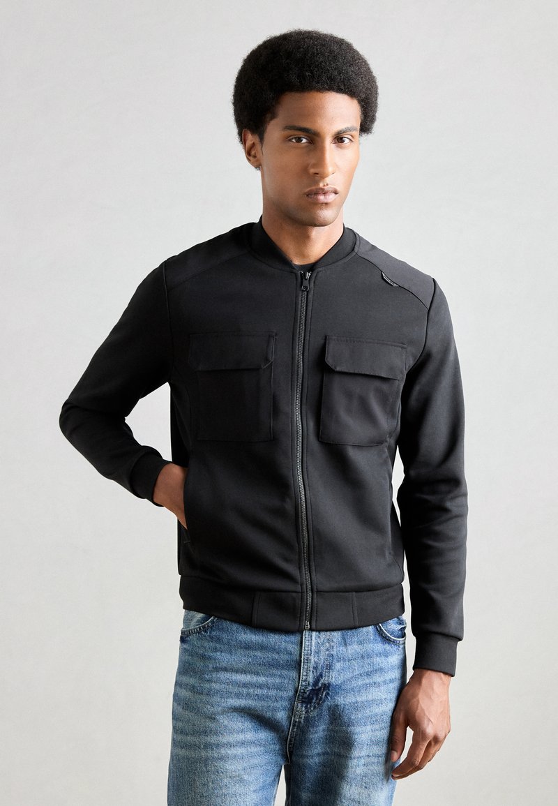 Veste bomber noire zippée avec deux poches poitrine, col et poignets côtelés, fabriquée en tissu lisse, associée à un jean en denim bleu clair.