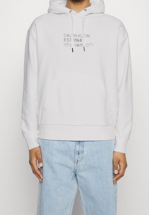 Biała bluza z kapturem z przednią kieszenią i szarym napisem "CALVIN KLEIN EST 1968 NEW YORK CITY", noszona z jasnoniebieskimi dżinsami.