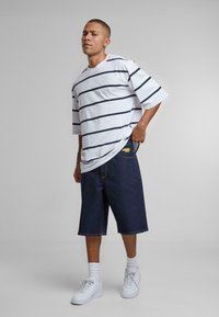Camiseta blanca a rayas con mangas cortas, pantalones cortos de mezclilla azul y zapatillas blancas. El modelo posa con una mano en la cadera, mostrando los detalles de la ropa.