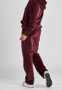 Burgunderfärgad velourträningsoverall med zip-up hoodie och byxor med elastisk midja; slät textur, avslappnad passform och sneakers med kontrasterande gummisula.