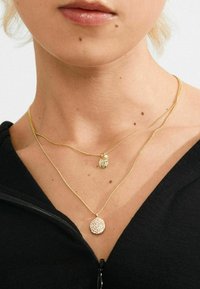 Gelaagde gouden kettingen met een getextureerde ronde hanger en een kleine versierde charm, rustend op een zwarte zip-up top.