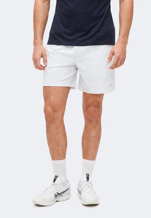 Mężczyzna w białych sportowych szortach, białych skarpetach do połowy łydki, białych butach do biegania i granatowej koszulce stoi na jednolitym tle.