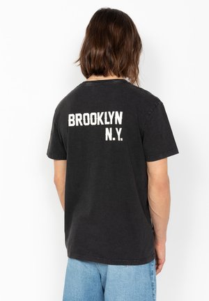 Persona con capelli castani fino alle spalle che indossa una maglietta nera con la scritta bianca "BROOKLYN N.Y." sulla schiena e jeans azzurri chiari.