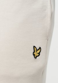 Tissu beige avec un petit logo oiseau brodé en or et noir près d'une couture verticale sur le côté droit.