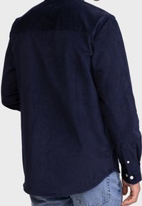 Marineblauwe corduroy shirt met lange mouwen, knoopmanchetten en een klassieke kraag; gladde textuur met subtiele glans en ton-sur-ton stikseldetails.