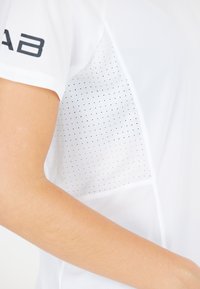 T-shirt de sport blanc avec un tissu texturé, des panneaux en maille et de petits points noirs. Manches courtes et logo sur le bras gauche.