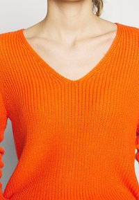 Stickad orange tröja med V-halsdesign, med strukturerad tyg och långa ärmar. Materialet verkar mjukt och andningsbart.