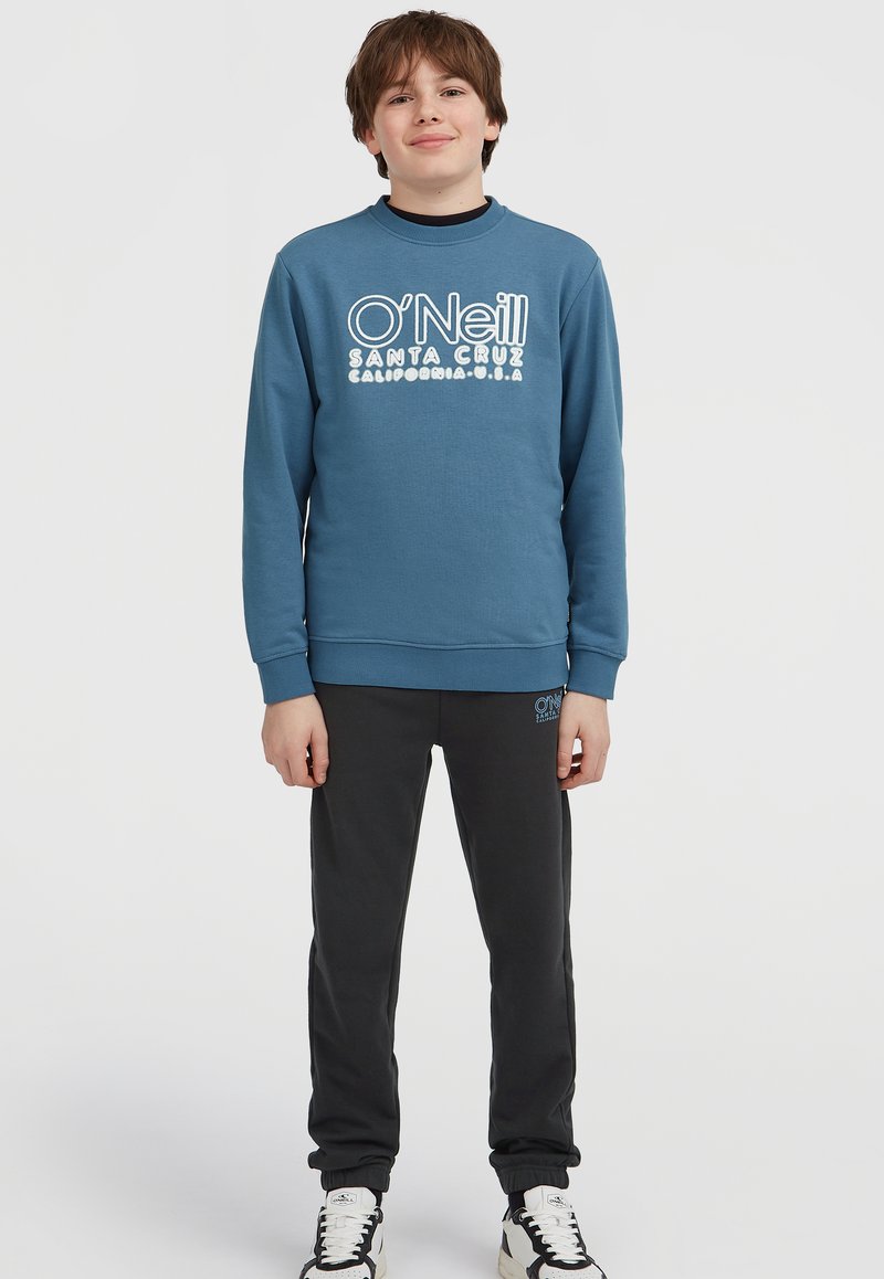 Blauwe crewneck sweatshirt met wit O'Neill-logo, gecombineerd met zwarte joggers en witte sneakers. De stof lijkt zacht en casual.