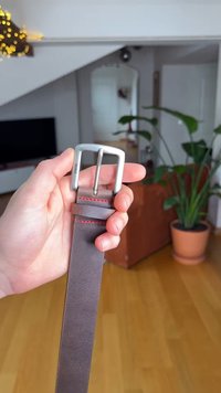 Brauner Ledergürtel mit einer silbernen Metallschnalle. Der Gürtel verfügt über rote Nähte und eine glatte Textur und wird in einer Hand gehalten.