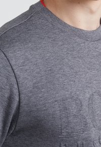 Gros plan sur une personne portant un pull en maille gris avec des lettres discrètement embossées et un petit détail rouge au niveau de l'encolure.