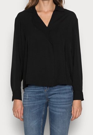 Žena v černé košili s dlouhým rukávem a límečkem a modrých vybledlých džínách, stojící před jednobarevným bílým pozadím.