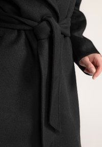 Zwarte jas van zacht, textuurstof. Beschikt over een strikceintuur in de taille en open mouwen. Close-up toont nette stiksels en elegante plooien.