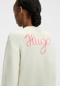 Biały sweter z dzianiny o luźnym kroju, z różowym, kursywnym logo „Hugo” na plecach. Miękka tekstura i okrągły dekolt.