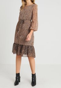 Robe imprimé léopard marron avec manches longues bouffantes et taille à cordon. Présente un ourlet à volants et est associée à des bottines noires.