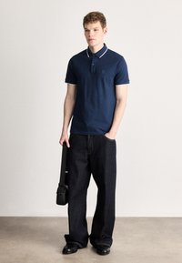 Tricou polo bleumarin cu guler alb, mâneci scurte și logo mic; asortat cu pantaloni evazați din denim închis și pantofi negri, ținând o geantă neagră.