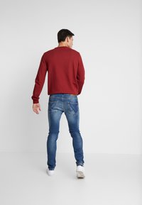 Punainen neulepaita pitkillä hihoilla, joka on puettu sinisten slim-fit farkkujen päälle. Farkuissa on klassinen viiden taskun muotoilu ja valkoiset lenkkarit.