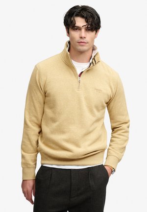 Jeune homme aux cheveux bouclés foncés portant un pull beige à fermeture éclair quart de zip et un pantalon sombre, debout avec une main dans la poche sur fond blanc.