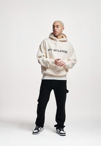 Cream hoodie with "2Y STUDIOS" text, paired with black cargo pants and black-and-white sneakers. Smooth fabric, relaxed fit, subtle design.