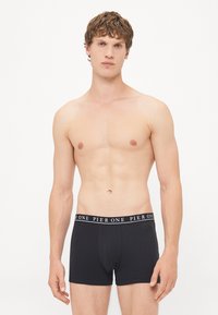 Schwarze Baumwoll-Boxershorts mit einem Logo-Bund, körperbetontem Design und glatter Textur, präsentiert an einem Modell vor einem schlichten Hintergrund.
