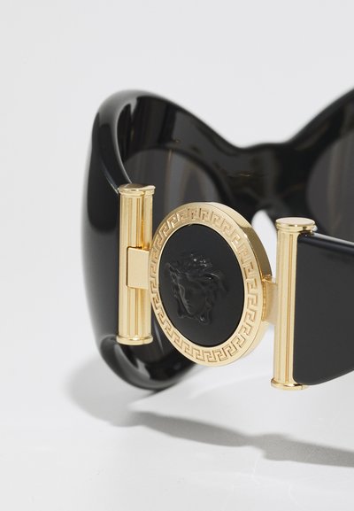 Lunettes de soleil noires surdimensionnées avec une finition brillante, dotées de quincaillerie dorée et d'un médaillon noir circulaire avec un design sculpté.