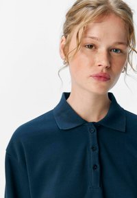 Polo shirt blu navy con colletto classico, cinque bottoni e tessuto strutturato. Il modello ha capelli ricci e orecchini in vista, volto in avanti.