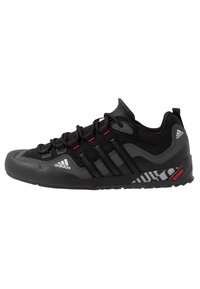 Chaussure de sport noire avec tige en mesh, embout en caoutchouc et semelle texturée. Présente trois rayures noires et des accents rouges. Logo sur le côté.