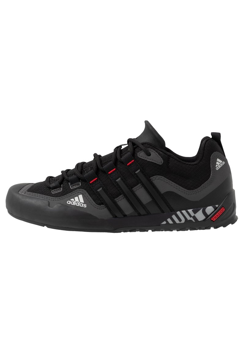 Chaussure de sport noire avec tige en mesh, embout en caoutchouc et semelle texturée. Présente trois rayures noires et des accents rouges. Logo sur le côté.