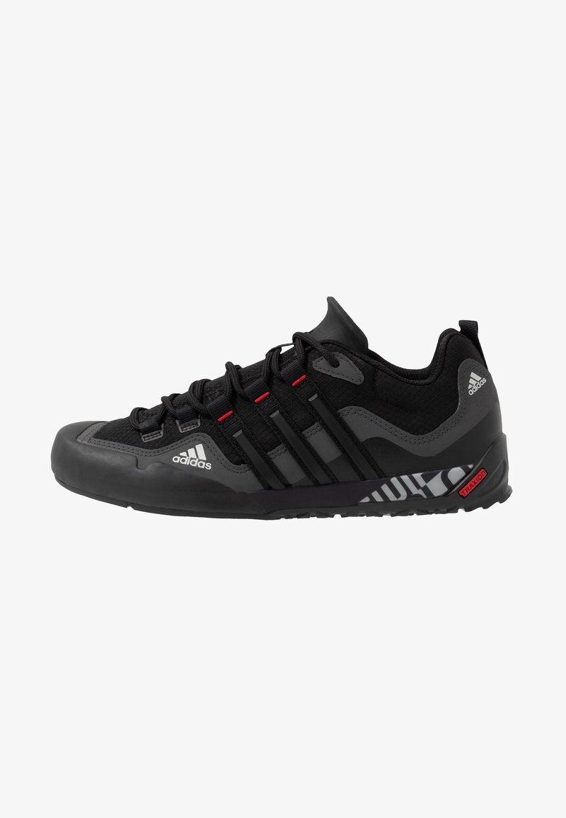 Chaussure de sport noire avec tige en mesh, embout en caoutchouc et semelle texturée. Présente trois rayures noires et des accents rouges. Logo sur le côté.