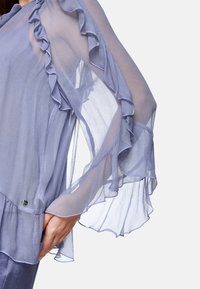 Lightweight, sheer lavender blouse with ruffled sleeves, textured fabric, and a decorative metal accent near the hem.