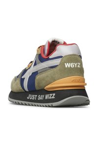 Zadní úhel vícerozměrného tenisky s brandingem W6YZ, béžovou semišovou kůží, modrou textilií, červenou podšívkou a nápisem "JUST SAY WIZZ" na černé podrážce.