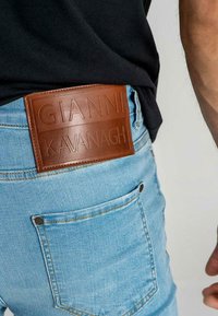 Acercamiento al bolsillo de unos vaqueros azul claros con una etiqueta de cuero marrón que dice "GIANNI KAVANAGH" en una persona que lleva una camiseta negra.