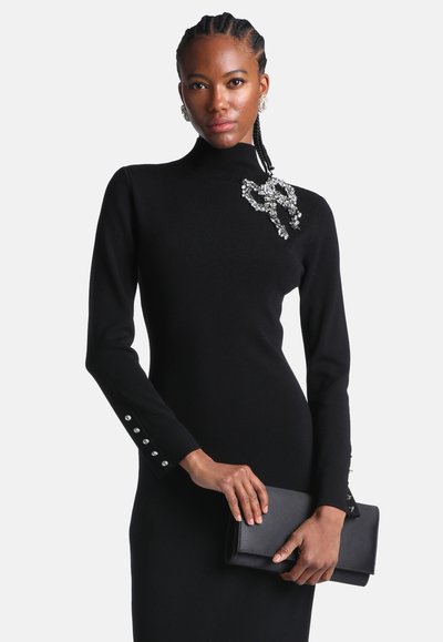 Femme portant une robe noire ajustée avec une broche en cristal, tenant une pochette noire et portant des boucles d'oreilles en cristal assorties avec les cheveux tressés.