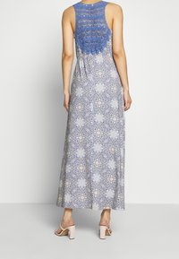 Une robe maxi avec un dos en crochet bleu, une jupe à motif floral dans des couleurs claires et une coupe décontractée. Assortie avec des sandales à talons blocs légers.