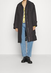 Manteau long noir avec capuche, comportant une fermeture éclair frontale, porté sur un cardigan jaune, associé à un jean bleu et des bottines noires.