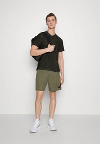 Camiseta preta de manga curta com textura subtil, calções verde-oliva com logótipo preto e ténis brancos. Modelo posa com uma mochila preta.