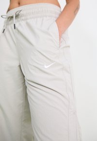 Světle šedé kalhoty Nike s elastickým pasem, bočními kapsami a bílým logem. Vyznačují se volným střihem a hladkým, lehkým materiálem.