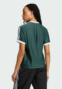 Zielony sportowy trykot z pionowymi paskami, białym kołnierzykiem i zdobieniami na rękawach. Model ubrany w czarne teksturowane spodnie, pokazany od tyłu.