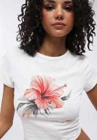 T-shirt bianca a maniche corte con un design di fiori hibiscus rosa e rossi, accompagnati da foglie verdi, realizzata in un morbido materiale di cotone.