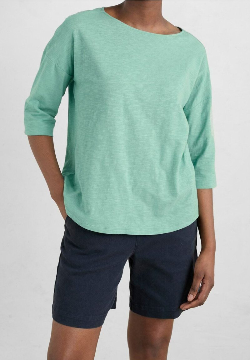 Personne portant un haut vert menthe à manches trois-quarts et un short bleu marine foncé, debout avec une main dans la poche et l'autre détendue à côté.