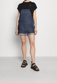 Džínové overalové šortky v tmavém odstínu s rolled cuffs, mají přední kapsy a nastavitelné popruhy. Vybavené černými sandály.