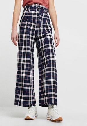 Personne portant un pantalon large à taille haute à carreaux bleu marine, blanc et rouge avec une ceinture, associé à des baskets blanches et un haut couleur rouille.