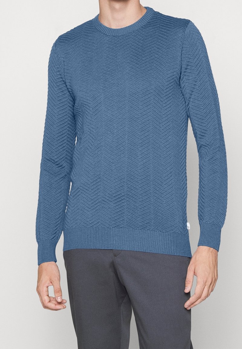 Pull en laine bleu tricoté avec un motif en chevrons texturé, col rond et poignets côtelés. Associé à un pantalon gris.