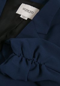 Blazer azul marino oscuro con una tela texturizada, que presenta un detalle de volantes en la manga y un forro negro liso. La etiqueta dice "SOAKED IN LUXURY."