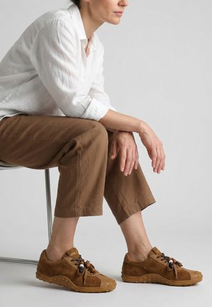 Femme portant un pantalon marron court et des chaussures marron lacées décontractées, assise sur un tabouret sur un fond gris uni.