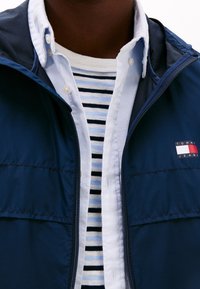 Tmavě modrá bunda na zip s logem, navrstvená přes bílo-modře pruhované tričko s bílým límečkem. Hladká textura, ležérní design.