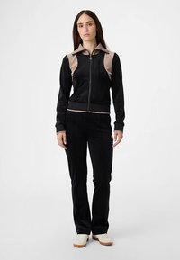 Schwarzer Velours-Trainingsanzug mit einer Reißverschlussjacke, die einen beigen Kragen und Brustpartien hat, kombiniert mit passenden Hosen und weißen Turnschuhen.